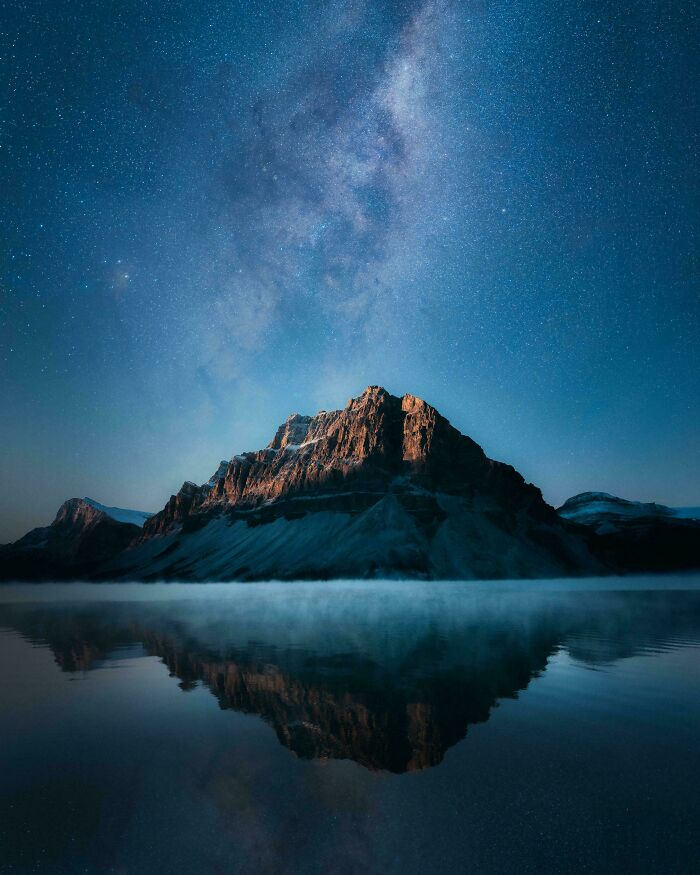 Mountain peak illuminated under starry night sky with Milky Way, reflected in calm water showcasing unique world perspective.