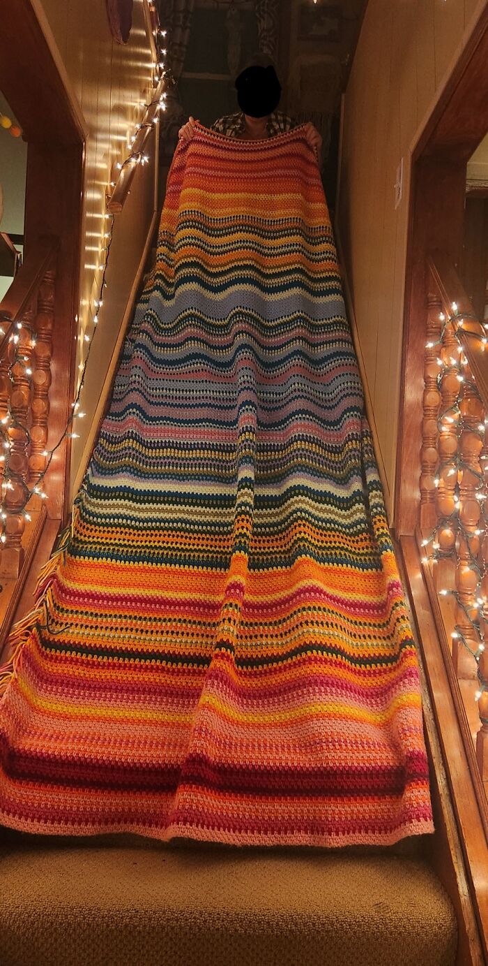 Person proudly holding a large colorful crochet blanket on stairs decorated with string lights, crochet enthusiasts project.