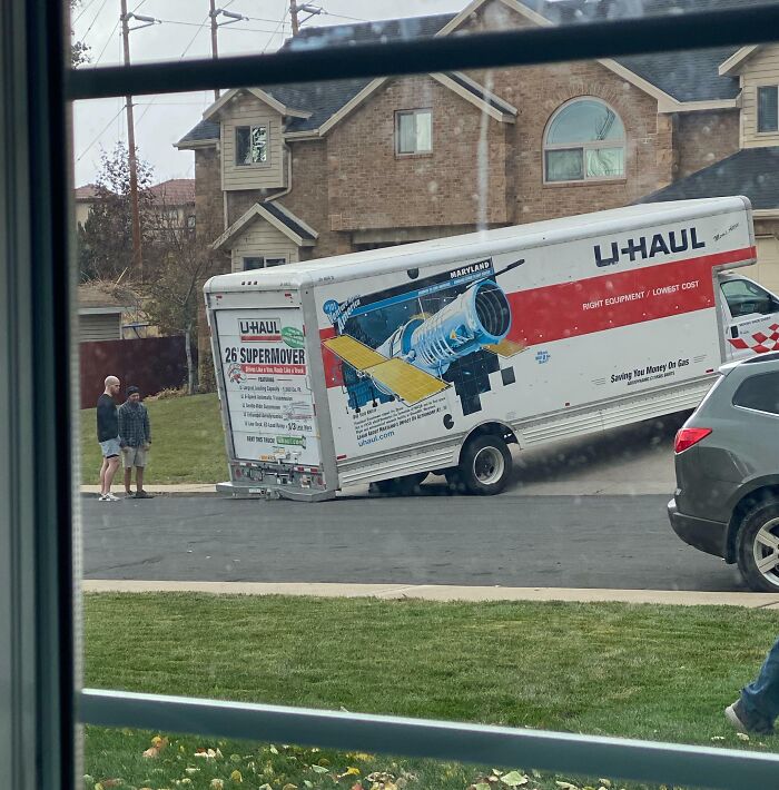 U-Haul truck stuck on its side in a neighborhood street as two people stand nearby, showing zero common sense.