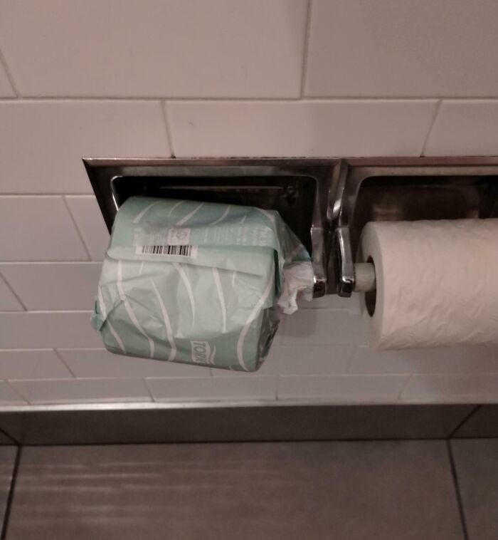 Toilet paper roll in packaging placed on holder next to regular roll, showing a lack of common sense in everyday situations.