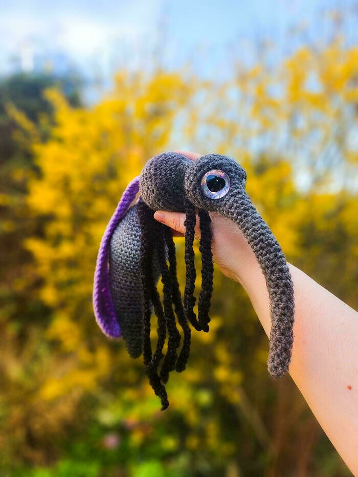 Hand holding a crocheted plush creature with large eye, long snout, and black braided tentacles in natural outdoor setting.
