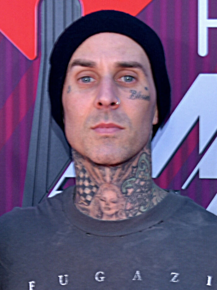 Travis Barker wearing a black beanie and tattooed neck in front of a colorful geometric background at an event.