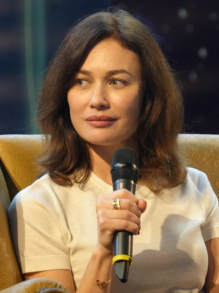 Olga Kurylenko speaking into a microphone during an interview, highlighting her bio and career achievements.