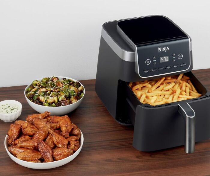 Ninja air fryer on wooden table with cooked fries, chicken wings, and a bowl of roasted vegetables, ideal gift for holiday shopping.