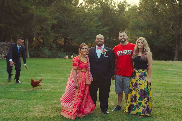 Group photo of four people smiling outdoors with an epic photobomb by a man chasing a chicken in the background.