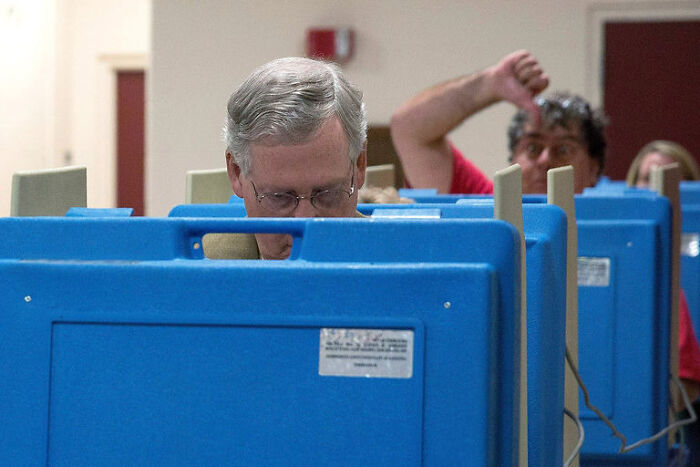 Man concentrating on voting behind booth while another person photobombs with thumbs down in epic photobombs collection.