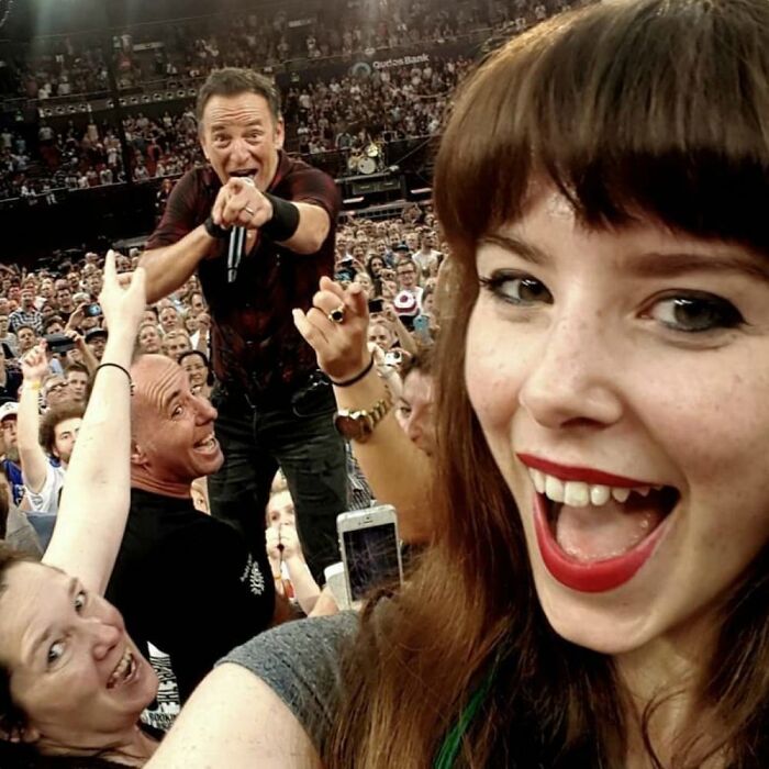 Young woman smiling for a selfie at a concert with a photobomb of a singer and excited crowd in the background.