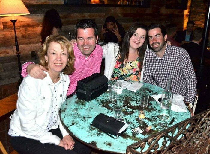 Group of four people smiling at a restaurant table with a photobombing figure in the background, capturing epic photobombs.