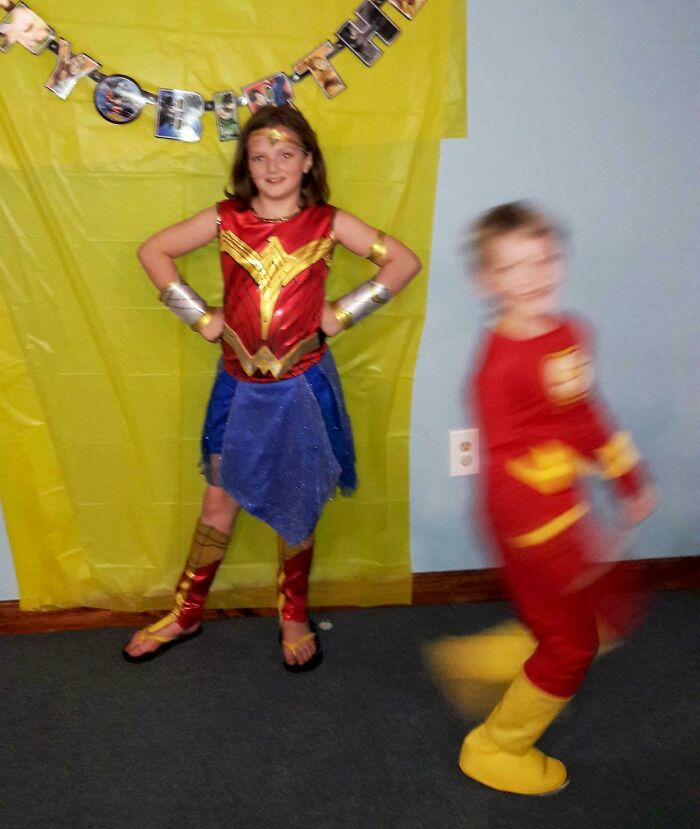Child dressed as Wonder Woman posing while a blurred child in a red costume photobombs, creating an epic photobomb moment.