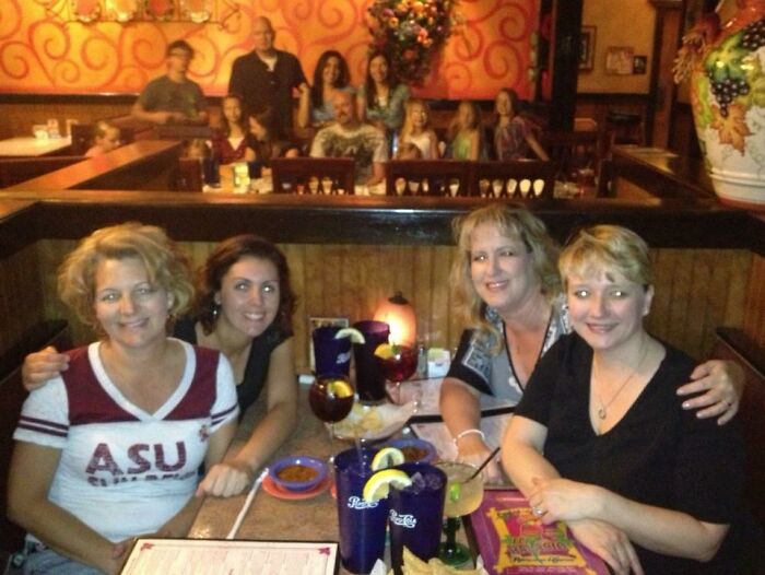 Group of women at a restaurant table with drinks, people in the background unintentionally photobombing the scene.