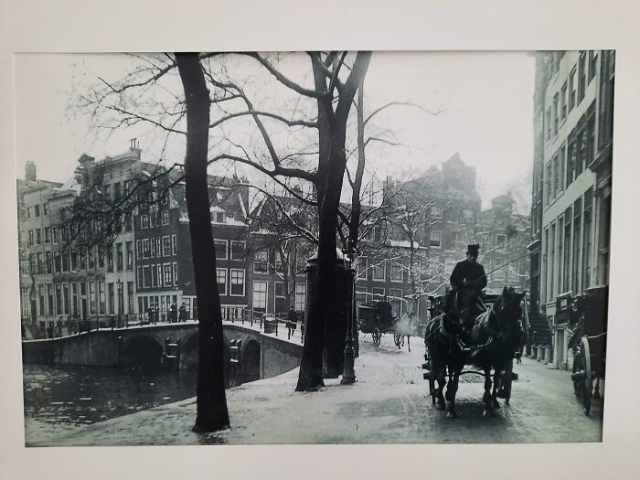 Black and white historic photo showing a snowy street, canal, and horse-drawn carriage in a European cityscape.