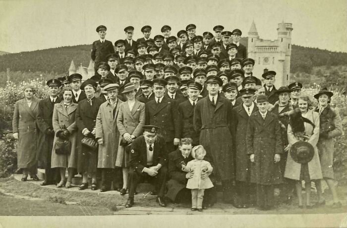 Large vintage group photo of people in uniform and coats outdoors with castle-like building in the background where was this taken