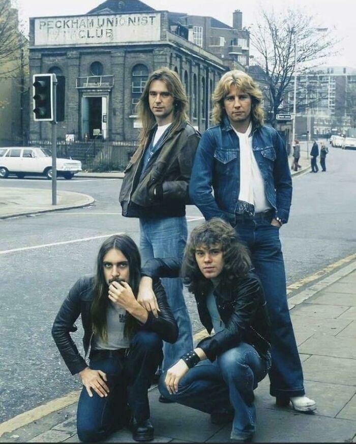 Four men in 70s-style clothing posing on a street corner near a building with a visible Peckham Unionist Club sign.