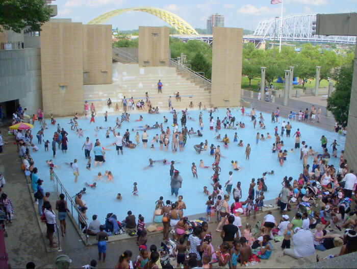 Crowded public pool with families and children playing near urban bridges and green spaces in a popular internet free time post.