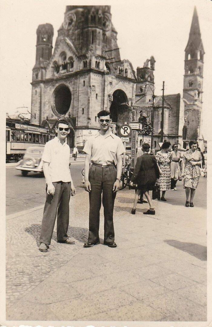 Two men wearing sunglasses posing on a city street with historic buildings and pedestrians in the background.