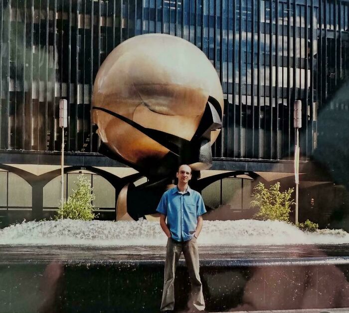 Man standing in front of the large iconic bronze sphere sculpture in an urban plaza, with water fountain and skyscraper backdrop.
