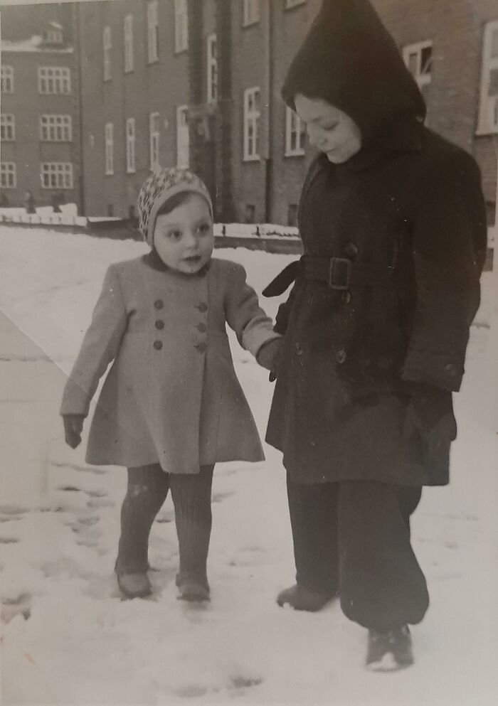 Two children holding hands walking in snow, wearing winter coats and hats, capturing a vintage where was this taken moment.