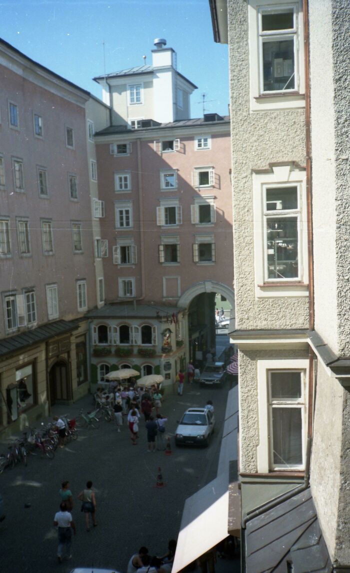 Narrow European street scene with people, bicycles, and outdoor cafe under clear blue sky in a historic town center.