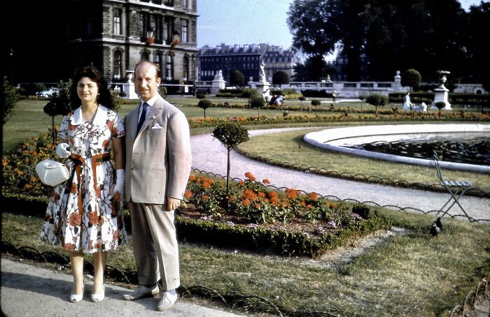 Couple dressed vintage style posing in a formal garden with flower beds and historic buildings in the background.