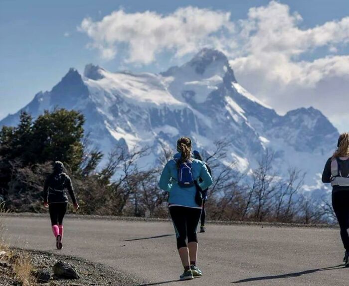 Hikers walking on a paved trail with snowy mountains and cloudy sky in the background in an outdoor setting.