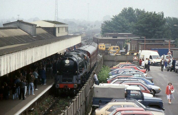 Vintage steam train arriving at a crowded station platform with parked cars and people walking nearby, where was this taken post.