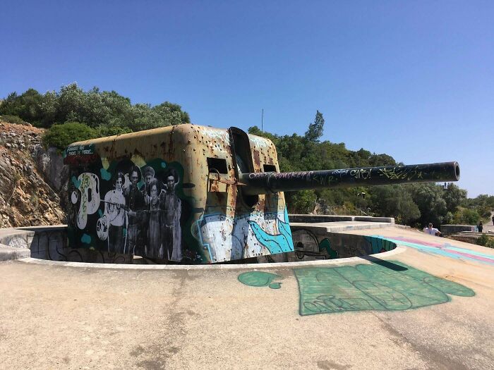 Rusty abandoned artillery with graffiti art on concrete platform, surrounded by trees under clear blue sky, where was this taken.