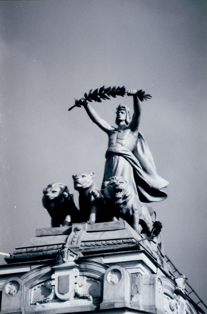 Black and white photo of a statue with a man holding a laurel wreath above three lions on a decorated building roof.
