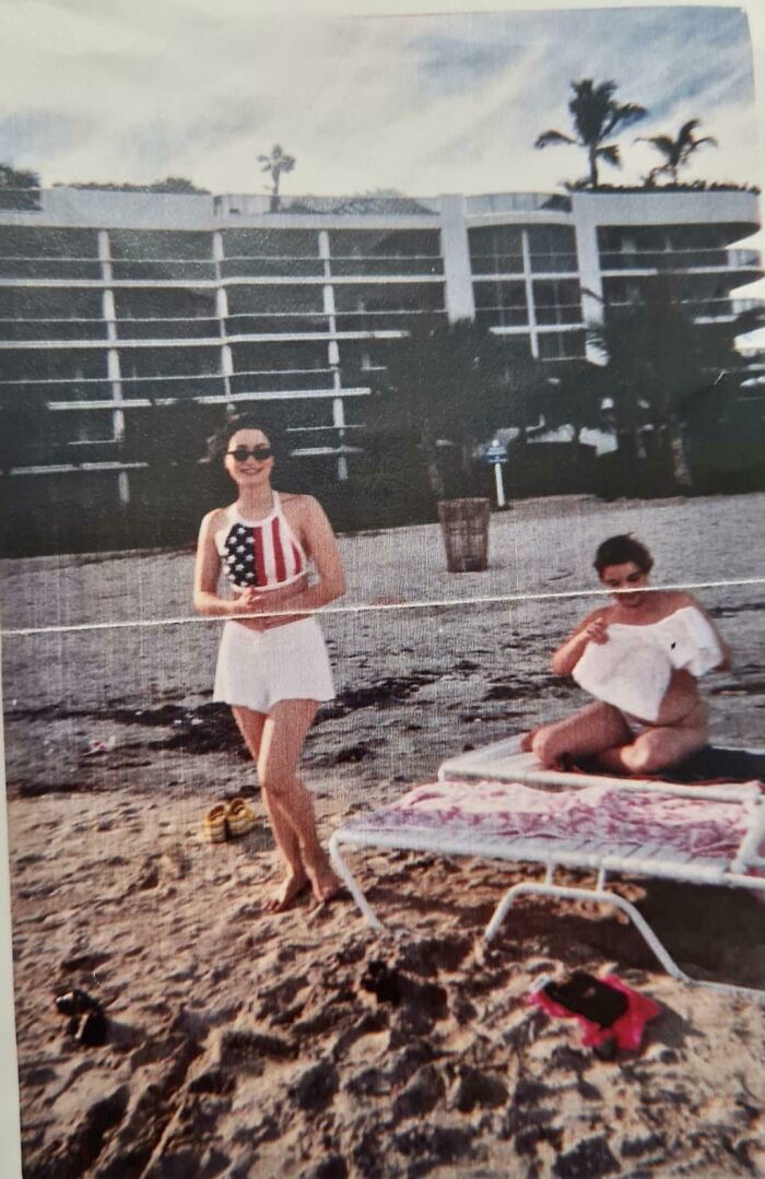 Two women on a sandy beach near a volleyball net with a white building and palm trees in the background, casual setting.