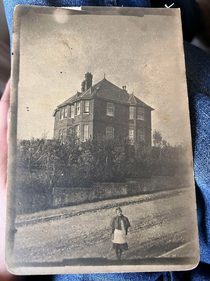 Vintage black and white photo of a large old house and a woman standing on a dirt road, mystery location guessing post.