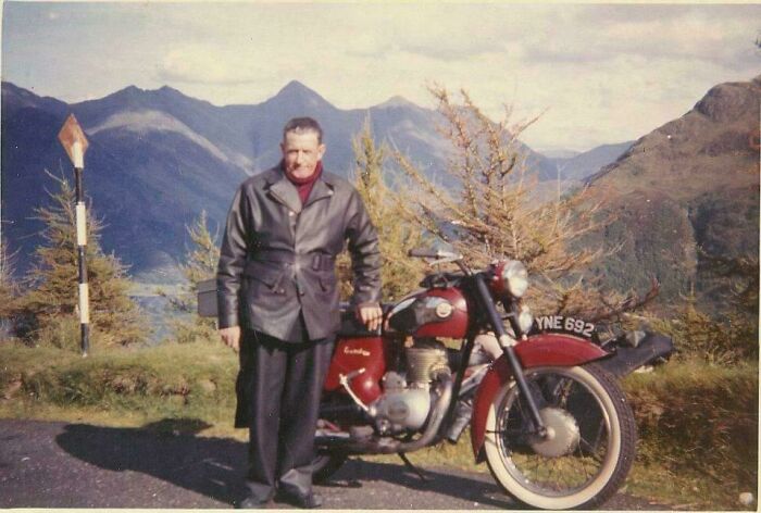 Man standing next to vintage motorcycle with mountainous background in a scenic outdoor setting from internet free time posts