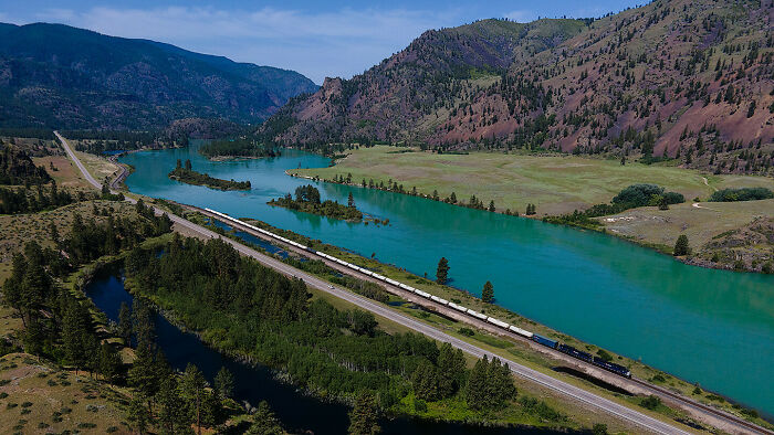 A long freight train traveling alongside a winding river in a mountainous landscape, showcasing nature and travel.