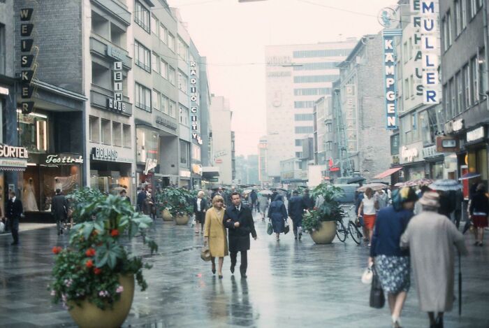 Busy city street with people walking past shops and planters on a rainy day in a popular where was this taken post.