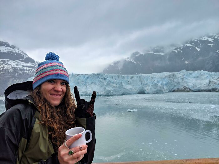 Woman wearing a colorful beanie holding a cup and smiling by a glacier, showcasing dreams turned into reality and inspiration.