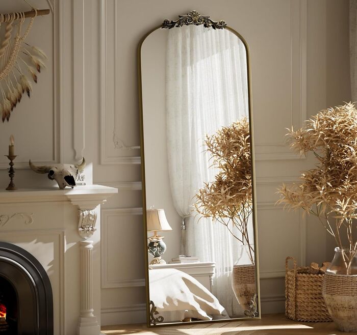 Tall ornate mirror reflecting soft natural light, beside dried plants and cozy fireplace in a warm inviting living space.