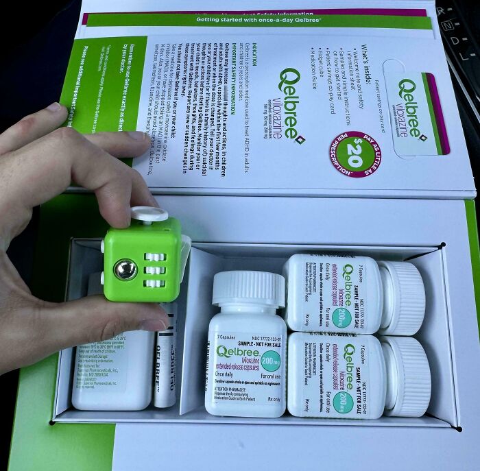 Hand holding green fidget cube inside a medication organizer with Qelbree pill bottles, showcasing smart ideas and inventions.