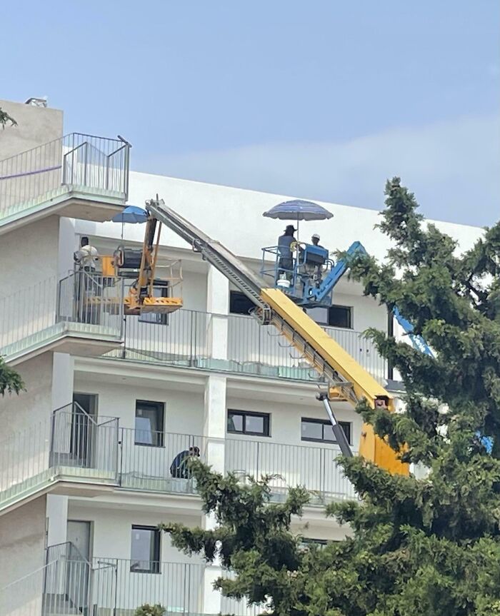 Construction workers using innovative lift equipment with built-in umbrellas on a building balcony for shade and safety.