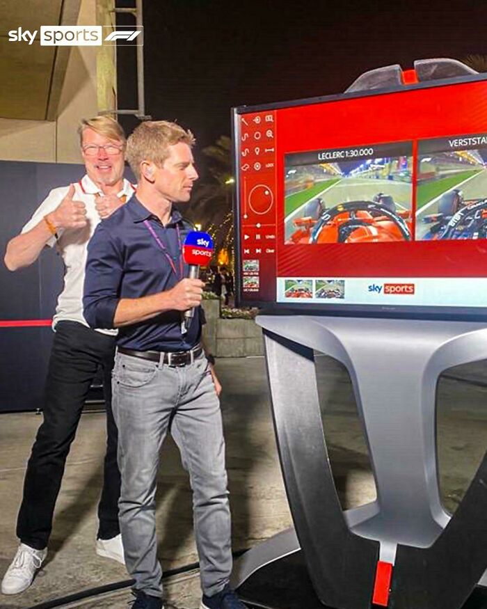 Formula 1 photobomb with a man giving thumbs up behind a reporter holding a Sky Sports microphone by a large screen.
