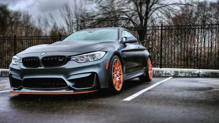 A sleek matte black BMW parked in an empty lot with bright orange wheels and accents under a cloudy sky.