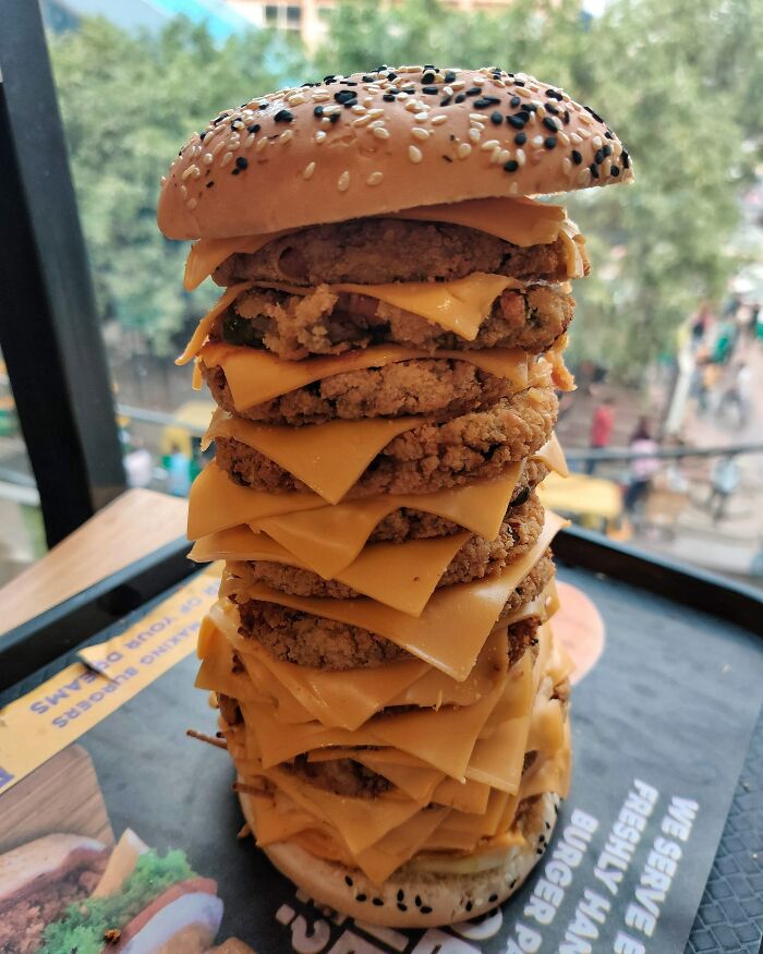 Stacked crispy fried patties with multiple slices of melted cheese in a tall burger bun topped with sesame seeds and black seeds.