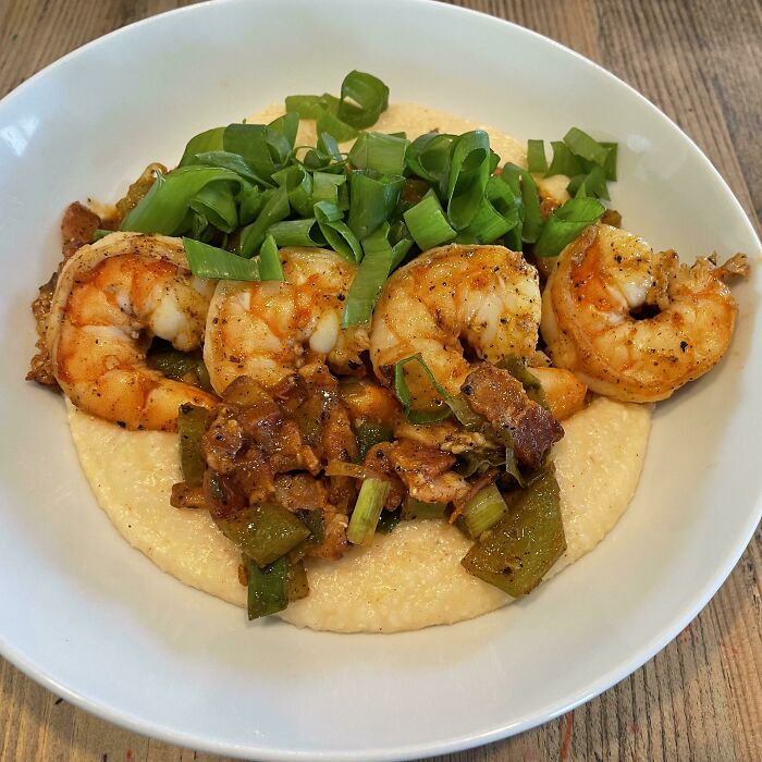 Plate of seasoned shrimp with vegetables and green onions served over creamy grits in a white bowl on a wooden table.