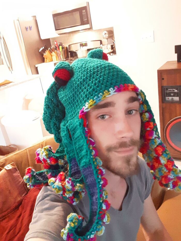 Young man wearing a colorful crocheted octopus hat, smiling in a cozy living room, showing sibling gratitude vibes.