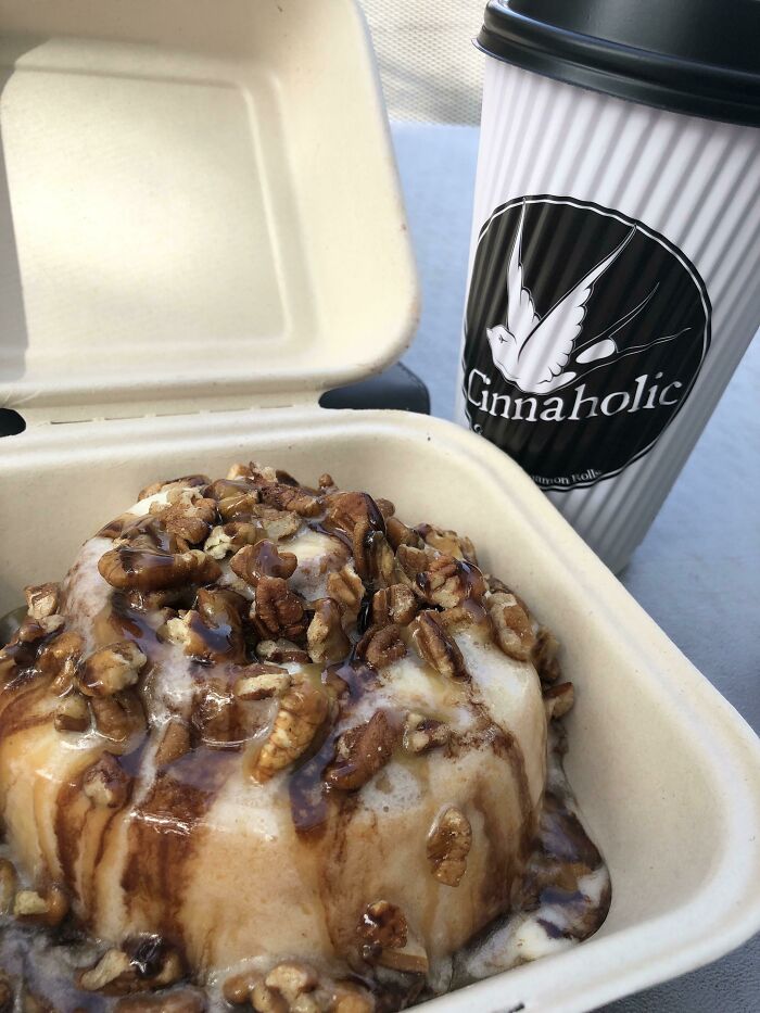 Cinnamon roll topped with nuts and glaze in a takeout container next to a Cinnaholic branded coffee cup on a table.