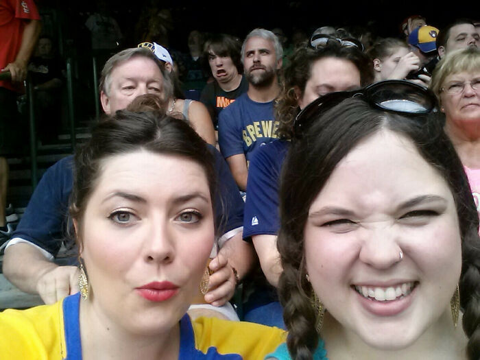 Two women taking a selfie at a crowded event with an epic photobomb in the background making funny faces.