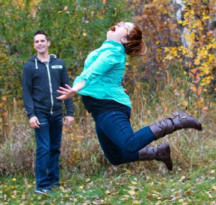 Woman in teal jacket jumping mid-air while man in hoodie smiles in background, an epic photobomb in autumn setting