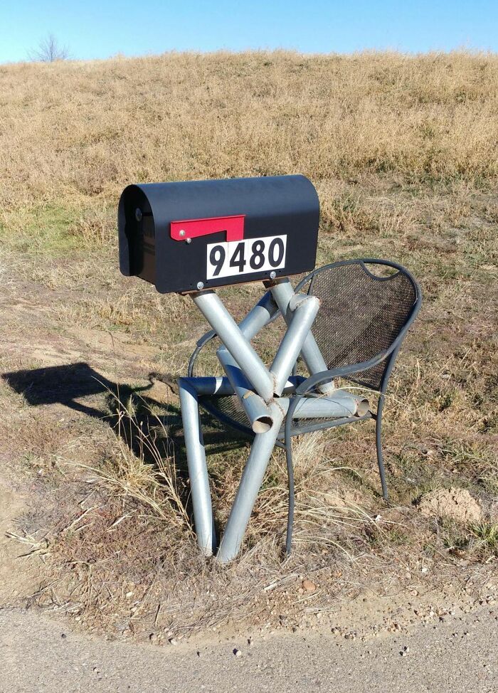 Creative and cool mailbox mounted on metal pipes next to a mesh chair along a rural roadside under clear sky.