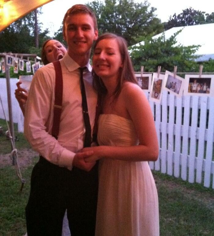 A couple posing for a photo with a funny epic photobomb behind them making everyone laugh outdoors.