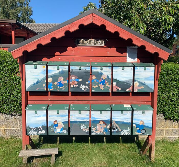 Group of creatively painted mailboxes under a red wooden shelter depicting playful children in a scenic outdoor setting