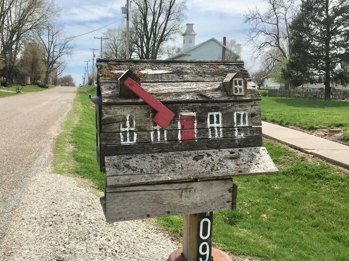 Creative and cool mailbox shaped like a rustic house with a red flag on a rural roadside in a green neighborhood.
