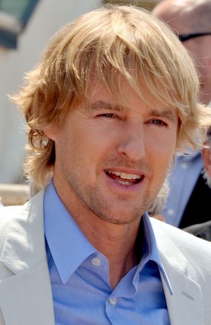 Owen Wilson smiling outdoors, wearing a light blue shirt and beige jacket, highlighting his bio and career in entertainment.