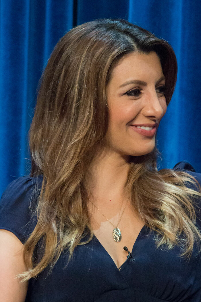 Nasim Pedrad smiling during an event, with long hair and wearing a dark blue top and gold pendant necklace.